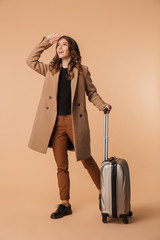 Wall Mural - Image of traveler woman 20s wearing stylish coat walking with baggage, isolated over beige background