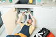 © Seventyfour - Positive handsome bearded plumber in uniform lying on flor and cutting cables while fixing water pipe in kitchen