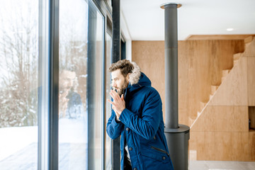 Man dressed in winter clothes feeling cold standing near the window at home with no heating