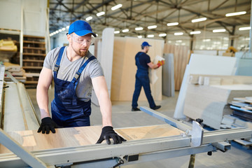 Serious concentrated handsome young worker in safety goggles standing at factory machine and measuring wooden piece while processing furniture