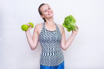 girl in a sports top holding a dumbbell and a bunch of green salad, the girl smiles