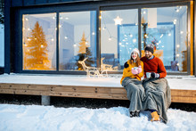 Young Couple Warming Up With Plaid And Hot Drinks Sitting On The Terrace Of The Modern House In The Mountains Durnig The Winter Holidays