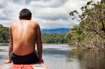 close up view from behind of young bare chested indigenous man with riding wooden boat in jungle river looking forward toward future