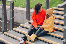 Image Of Fashionable Young Woman Wears Red Looe Sweater, Looks Positively At Small Child, Pose Together At Steps Outdoor, Enjoy Spare Time, Stroll In Urban Setting. People, Relationship Concept