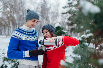 Young couple pouring hot tea out of thermos in winter forest. Happy people relaxing outdoors during holidays