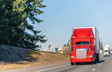 Big rig bright red classic American idol semi truck with reefer semi trailer driving on the straight highway in sunny day