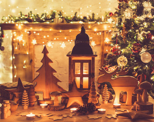 Wall Mural - Wooden toys, figures, stars, moose, fur tree lantern on table in front of fireplace, decorated mantel, Christmas tree with baubles, ornaments garlands, lights candles, selective focus, toned