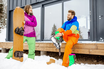 Young couple in ski suits preparing for snowboarding sitting on the terrace of the modern house in the mountains