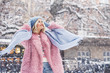 Outdoor portrait of young beautiful fashionable happy smiling girl wearing trendy pink faux fur winter coat, light blue beanie hat, scarf, posing in snow covered street. Copy, empty space for text