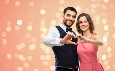 Wall Mural - valentines day, love and people concept - happy couple in party clothes making hand heart gesture over living coral background and festive lights