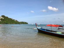 Boat On The Beach. Traditional Thai Longtailboat For Fishing With Red Roof, Anchored At The Shallow Water. Seascape: Green Cliff Of Island Extends To The Sea, Cloudy Sky, Horizon Line, Longtail Boat