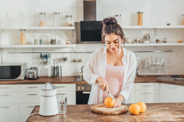 pretty young woman cutting orange by knife on wooden board in kitchen