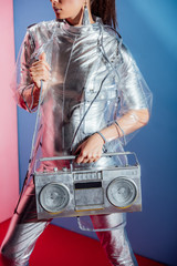 cropped view of girl in silver bodysuit and raincoat posing with boombox on pink and blue background