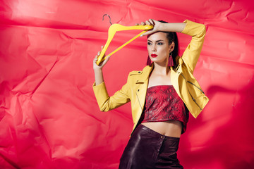 Wall Mural - beautiful stylish model in yellow leather jacket posing with hanger on red background