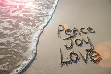 Happy Holiday Message Of Peace, Joy, And Love Handwritten In Smooth Sand With An Oncoming Wave In The Lens Flare Of The Tropical Sun