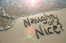 Cheeky Christmas Message Naughty Or Nice? Handwritten On Smooth Sand Beach With Incoming Wave