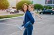 Image of affectionate mother gives piggyback to small daughter, smile broadly, have outdoor walk, pose against blurred background with cars, play together. People, family and happiness concept