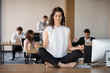 © fizkes - While diverse colleagues working, attractive young employee in coworking area sitting on office table in lotus pose and practising yoga meditating folding fingers in Jnana Mudra. Stress relief concept