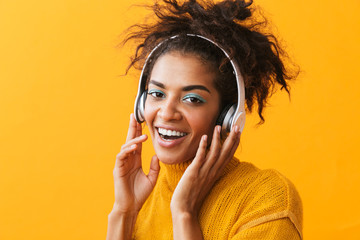 Poster - African american glad woman in casual clothing listening to music via white headphones, isolated over yellow background