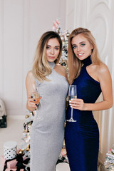 Incredible two blond women in elegant evening dresses with champagne on background of Christmas tree celebratinig new year, Christmas. Two friends on birthday party