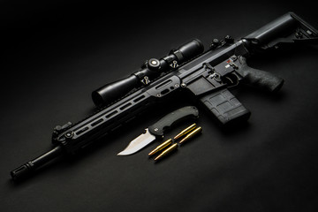 modern automatic rifle with an optical sight lies on a dark background and next to the knife and ammunition