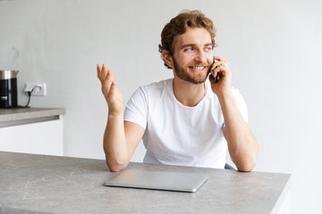 Wall Mural - Handsome young bearded man at the table at home talking by phone near laptop computer.