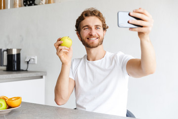 Wall Mural - Happy young bearded man at the table at home make a selfie by phone with fruits.