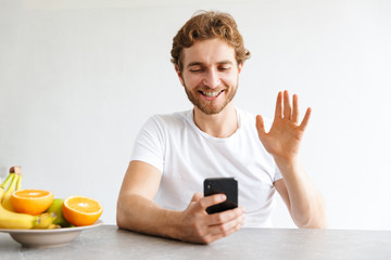 Wall Mural - Happy young bearded man at the table at home talking by mobile phone waving.