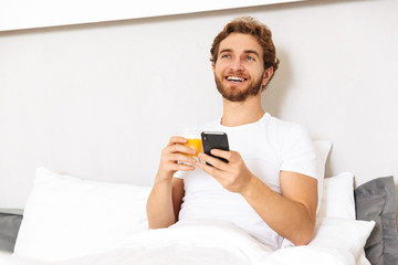 Wall Mural - Handsome young bearded man in bed at home drinking juice using mobile phone.