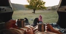 Beautiful couple holding hands in a car trunk and have a romantic time together wearing a cozy socks in the middle of nature drinking tea from iron cup. 4k