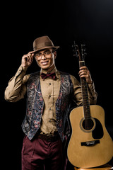 Wall Mural - cheerful mixed race male musician in eyeglasses and hat posing with acoustic guitar isolated on black