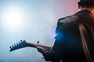 Wall Mural - rear view of male rock star in leather jacket performing on electric guitar on stage with smoke and dramatic lighting