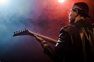 Wall Mural - rear view of male musician in leather jacket playing on electric guitar on stage with smoke and dramatic lighting