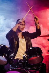 Wall Mural - mixed race male musician raising drum sticks while sitting behind drum set on stage with dramatic lighting and smoke