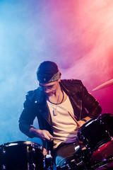 Wall Mural - concentrated young male musician in leather jacket playing drums during rock concert on stage with smoke and dramatic lighting