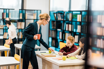 young female teacher holding cup and looking at schoolgirls studying in library