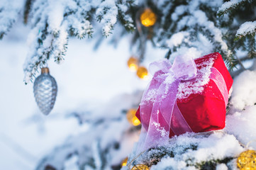 Close-up red New Year's gift with a white ribbon next to Christmas toys on the branches of a snow-covered Christmas tree in the winter forest. The concept of Christmas gifts and the new year