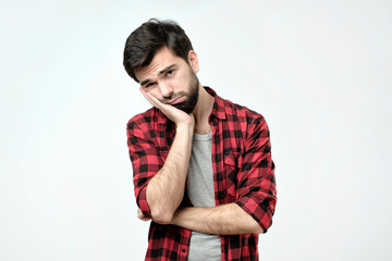 Very bored adult man with beard holding hand on cheek while support it with another crossed hand, looking tired and sick