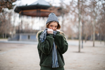 Pretty young female in warm clothes wrapping in scarf and looking away while standing on blurred background of autumn park on really cold day