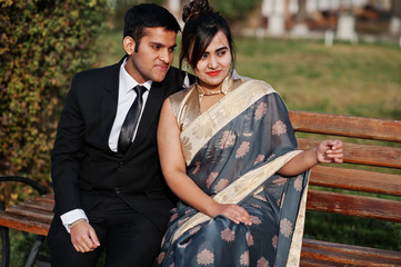 Wall Mural - Elegant and fashionable indian friends couple of woman in saree and man in suit sitting on bench.