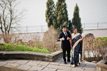 Wall Mural - Elegant and fashionable indian friends couple of woman in saree and man in suit posed on stairs against lake.