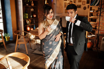 Wall Mural - Elegant and fashionable indian friends couple of woman in saree and man in suit sitting on cafe and drinking tea.