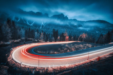 Blurred Car Headlights On Winding Road In Mountains With Low Clouds At Night In Autumn. Moody Landscape With Asphalt Road, Light Trails, Foggy Forest, Rocks And Blue Sky At Dusk. Roadway In Italy