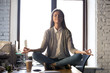 Serene calm business woman sit on office desk taking break for meditation, mindful employee doing yoga exercise in lotus pose for relaxation at workplace, no stress relief, balance at work concept