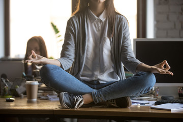 Young woman office worker student sitting on desk in lotus position doing yoga exercise for balance concentration at workplace, meditation and yoga at work for no stress relief concept, close up view