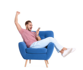 Wall Mural - Emotional young man with smartphone in armchair on white background