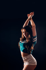 Wall Mural - Portrait of sexy attractive hot brunette woman with long wavy hair in short white shorts and blue t-shirt in the dark on black background. Slim figure, fitness, sports, dancing, twerk, hobby