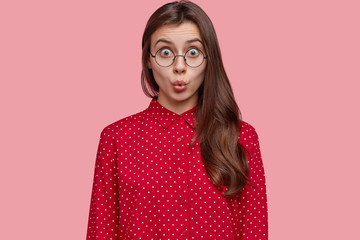 Poster - Photo of shocked woman in stupor, keeps lips round, has distrubing situation, dressed in fashionable polka dot blouse, wears round spectacles, expresses surprisement, isolated over pink wall