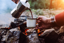 Tourists A Sits Near Camp Fire An In Summer Time And Pours Itself Hot Coffee During The Sunset . Concept Adventure Active Vacations Outdoor. Summer Camping.