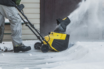 A man in a black jacket and a gray pants is brushing white snow with the yellow electric snow thrower in winter